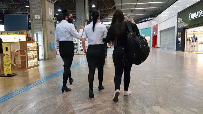 Mylena Rodrigues caminha pelo Aeroporto de Guarulhos, acompanhado de duas mulheres que estão prestando assistência. Mylena segura no braço de uma delas para ser conduzida.