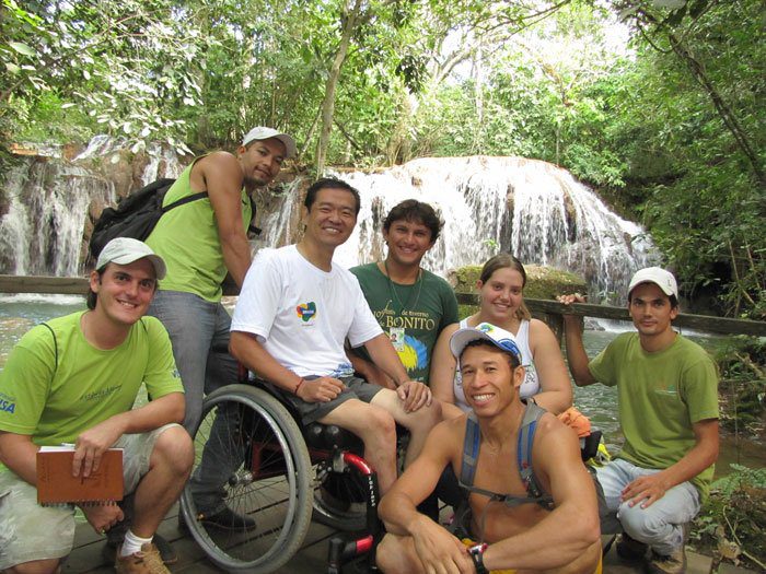 Ricardo Shimosakai está sentado em sua cadeira de rodas, acompanhado de mais 6 pessoas, em cima de um deck de madeira. Ao fundo aparece uma cachoeira e vegetação nativa de da cidade de Bonito.