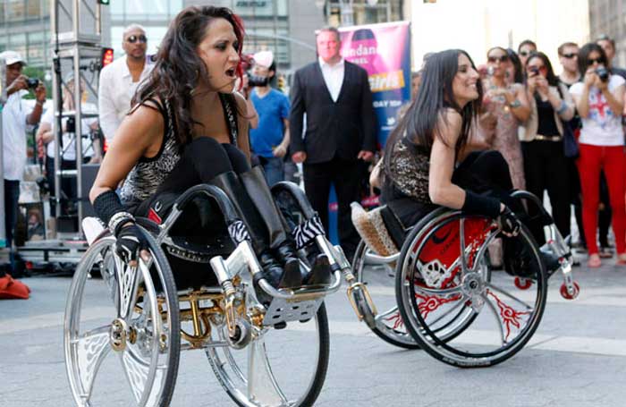 Duas garotas cadeirantes fazem manobras empinando a cadeira de rodas, assistidas por um grupo de pessoas