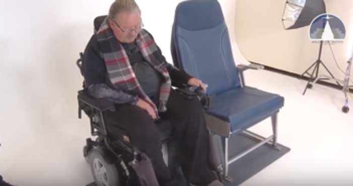 um senhor sentado em uma cadeira motorizada, está posicionado ao lado de uma poltrona de avião, num ambiente simulado de um estúdio fotográfico