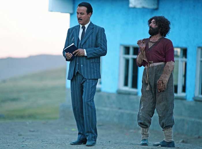 dois atores, na esquerda com terno e na direita com roupas rasgadas e sujas. Estão no quintal gramado de uma casa azul que aparece ao fundo.