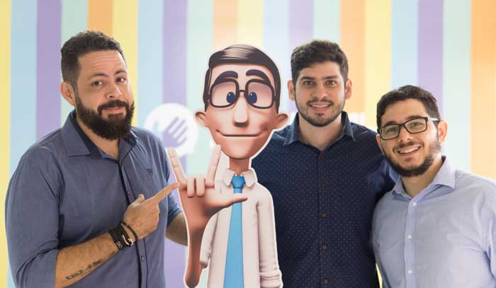 Os três criadors da Hand Talk estão de pé, ao lado de um boneco do assistente virtual Hugo. Ao fundo, papel de parede listrado na horizontal com diversas cores.