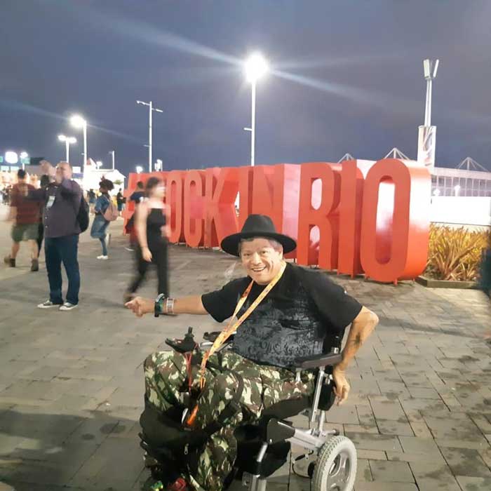 Homem em uma cadeira motorizada, em uma praça com a escultura da palavra Rock In Rio em vermelho atrás
