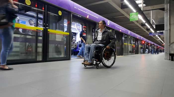 Acessibilidade em Destinos Turísticos Inteligentes. Foto com Ricardo Shimosakai sentado em sua cadeira de rodas na plataforma da Estação Adolfo Pinheiro, da linha Lilás do Metrô de São Paulo. Mostra também os vagões parados, de portas abertas com pessoas saindo deles.